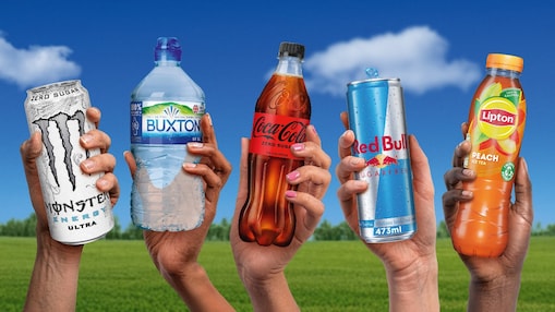 Five hands are holding different drinks against a bright blue sky with clouds and a green field. From left to right, the drinks are a white can of Monster Energy Ultra with “zero sugar” text, a Buxton still water bottle, a Coca-Cola Zero Sugar plastic bottle, a silver and blue Red Bull sugarfree can (473 ml), and a Lipton peach iced tea bottle labelled “low in calories”.