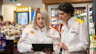 Two Shell employees in uniform collaborate with a tablet in a store, surrounded by colourful displays