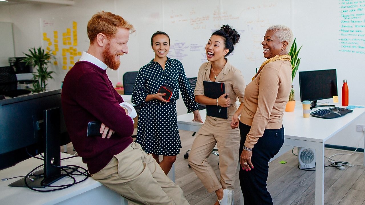 A diverse group of people in a modern office stand smiling and laughing by desks, fostering a cheerful and collaborative workplace