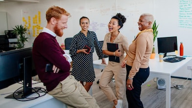 A diverse group of people in a modern office stand smiling and laughing by desks, fostering a cheerful and collaborative workplace
