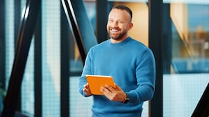 A smiling man in a blue sweater holds an orange tablet in a modern office with large windows
