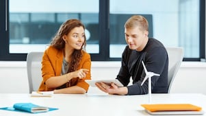  Two people discuss at a desk - one, wearing an orange blazer, points to a tablet held by the other