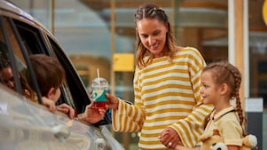 A woman in a yellow striped sweater hands a drink to a child inside a car with another child standing nearby holding a teddy bear