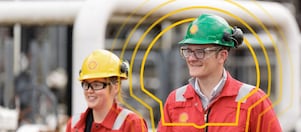 Two industrial workers wearing red uniforms and hard hats walk confidently amidst piping