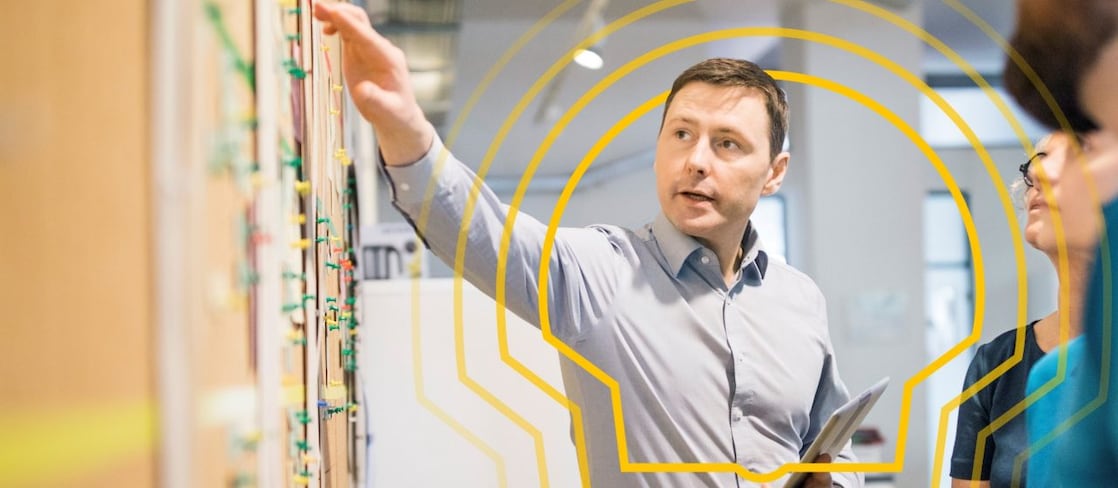 A man in a light shirt points at a wall map with various coloured pins, holding a tablet