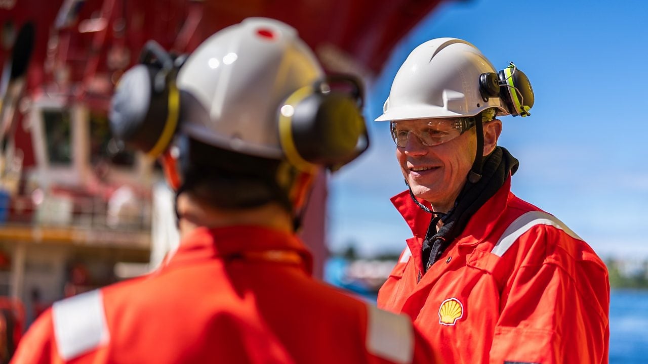 A worker in red coveralls with a Shell logo talks to a colleague.