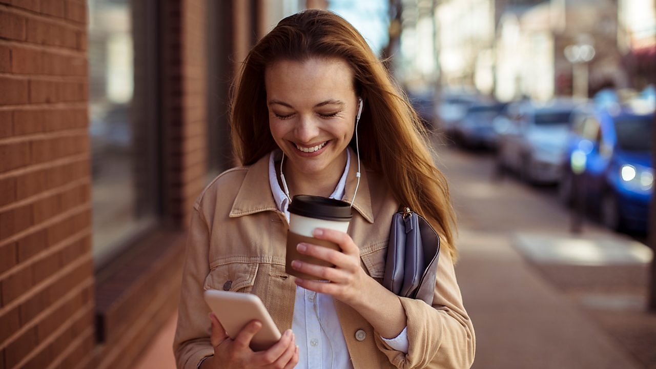 A smiling woman walks outdoors, holding a coffee cup