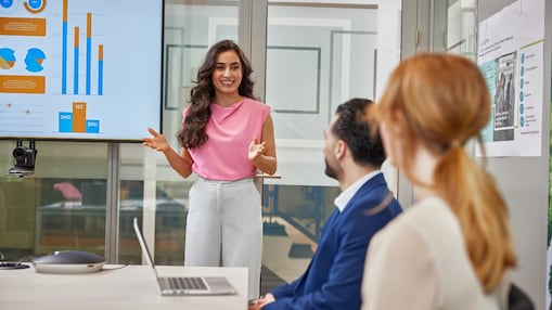 A woman in a pink blouse presents to two colleagues, gesturing toward a screen displaying colourful charts and graphs.