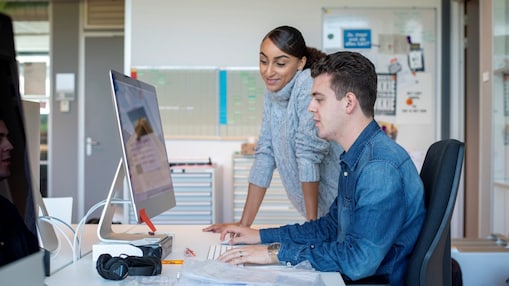 A woman in a grey sweater leans over to assist a man in a denim shirt working at a computer in an office
