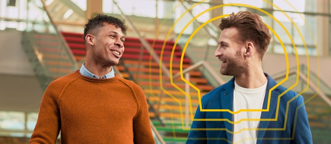 Two smiling men walk indoors with rainbow-coloured stairs in the background