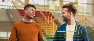 Two smiling men walk indoors with rainbow-coloured stairs in the background
