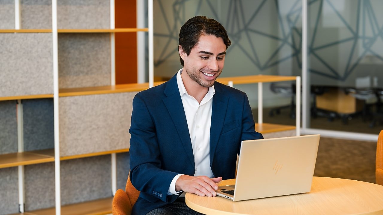 A man in a blue blazer smiles while working on a laptop at a wooden desk in a modern office