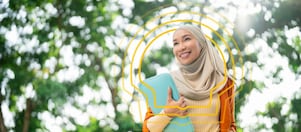 A woman wearing a beige hijab and orange blazer smiles while holding a blue folder outdoors