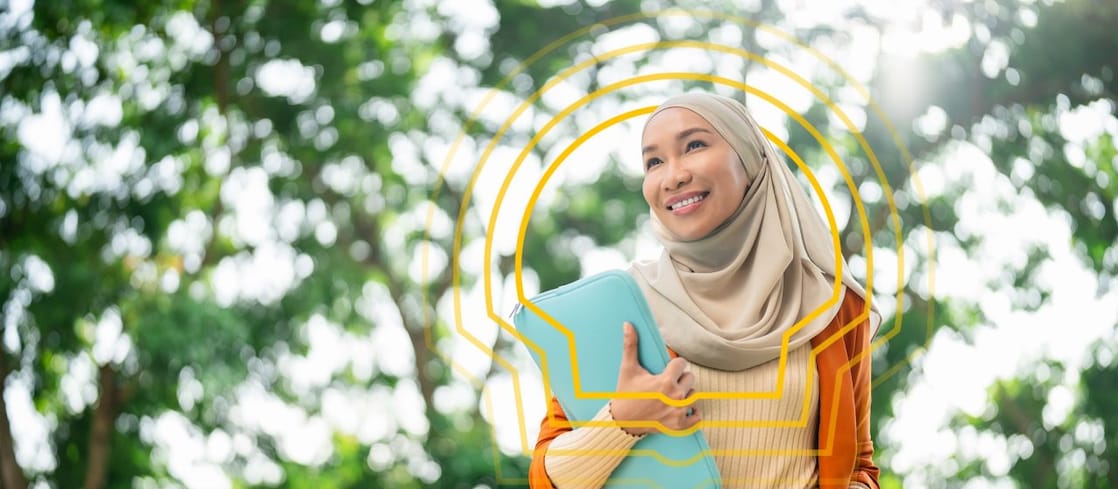 A woman wearing a beige hijab and orange blazer smiles while holding a blue folder outdoors