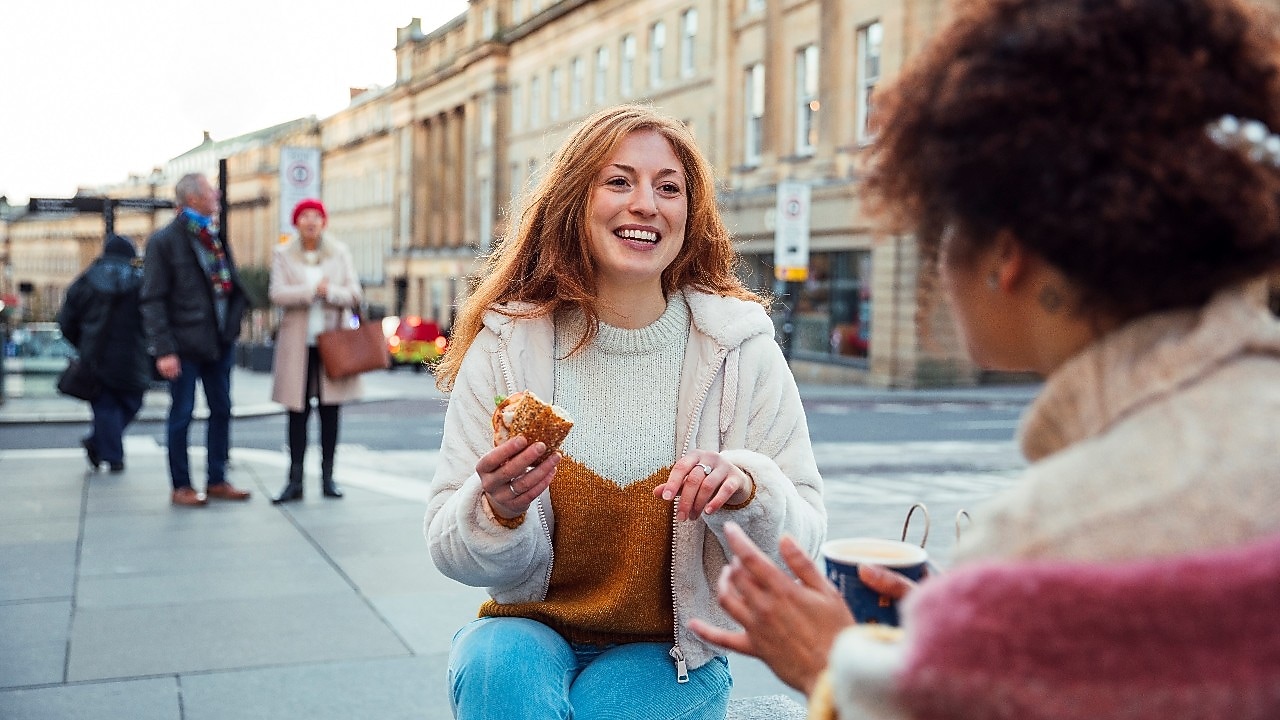 A woman with red hair, wearing a sweater, sits outdoors eating a sandwich, smiling at a friend holding a cup