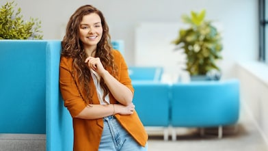  Smiling woman with curly hair leans on a blue panel, wearing an orange blazer and jeans