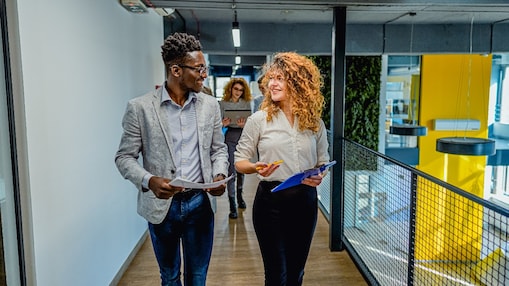 A smiling man and woman walk in a bright office hallway holding documents