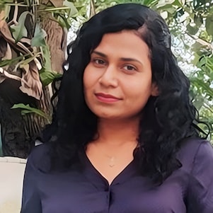 A woman with long black hair and a dark top stands smiling in a garden