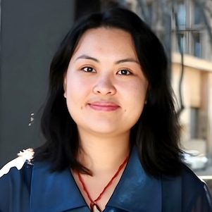 A woman in a blue shirt smiles softly with a blurred urban scene in the background