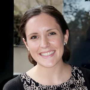 Smiling woman with dark hair tied back, wearing a patterned top, stands indoors with trees visible through the glass behind her
