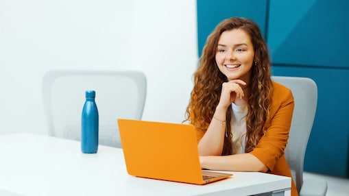 Woman is seated at a desk in a modern office setting, wearing an blazer and using an laptop in front of her. A reusable water bottle is placed on the desk. The background features geometric wall panels and a office chair.