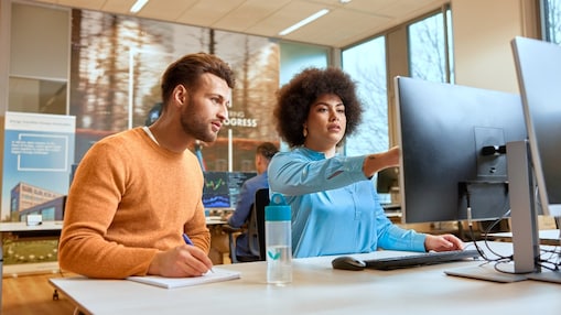 Two colleagues in an office, one pointing at a computer screen while the other takes notes
