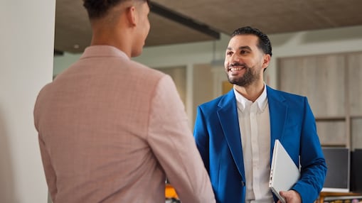 Two men in an office setting shake hands, smiling