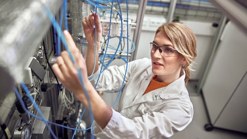A woman in safety glasses and a white lab coat works intently with blue cables in a laboratory