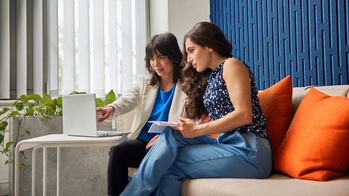 Two women sit on a couch in a modern office, engaged in discussion over a laptop