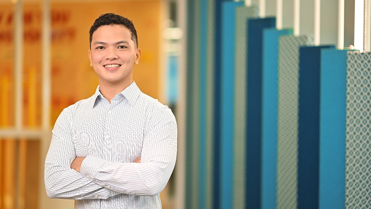 Smiling person with short curly hair in a light striped shirt stands with arms crossed in a modern office