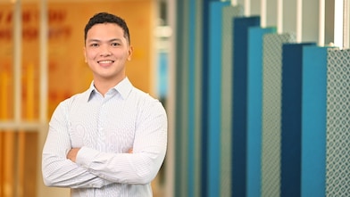 Smiling person with short curly hair in a light striped shirt stands with arms crossed in a modern office