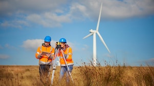 Two engineers in blue helmets and orange vests survey a field with equipment with a wind turbine in the background
