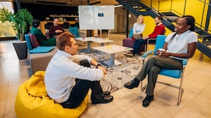 A man in a white sweater sits on a yellow bean bag facing a woman in a chair with colleagues in the background
