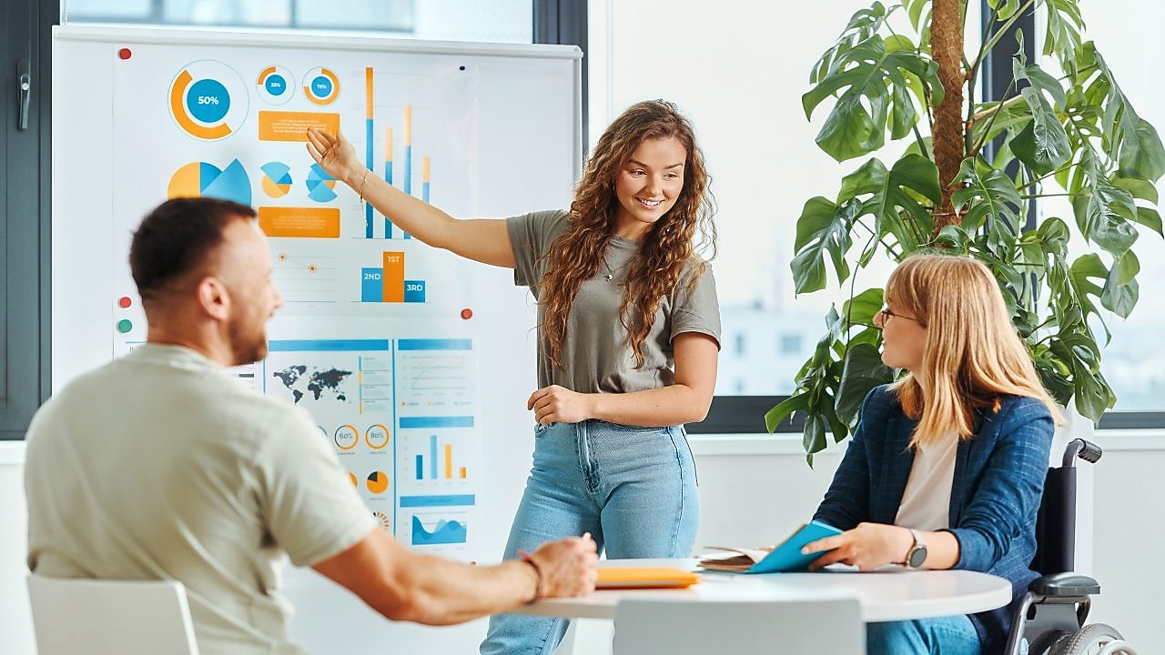 Young woman presenting charts on white board in front of man and woman sitting at the table.