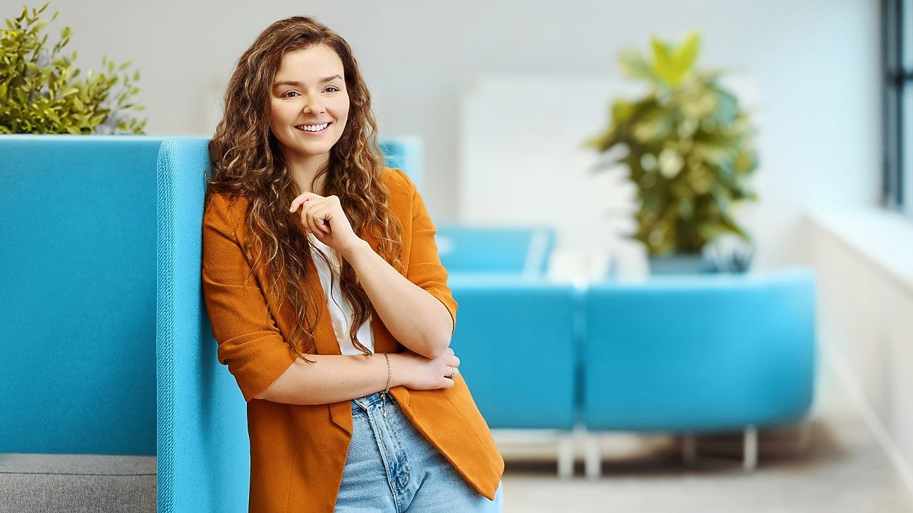  Smiling woman with curly hair leans on a blue panel, wearing an orange blazer and jeans