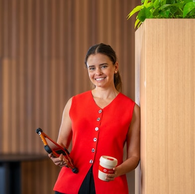 Young woman in red west standing and smiling, holding tablet and coffee mug.