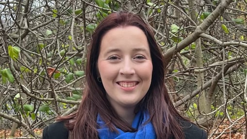 Front portret of young woman smiling, in medium dark hair and black jacket, with trees behind her.