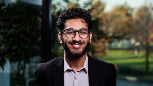 Front portret of a young man in a shirt and jacket wearing glasses and smiling.