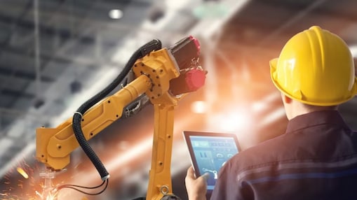 A worker in a hard hat is manipulating a factory robot arm with a tablet