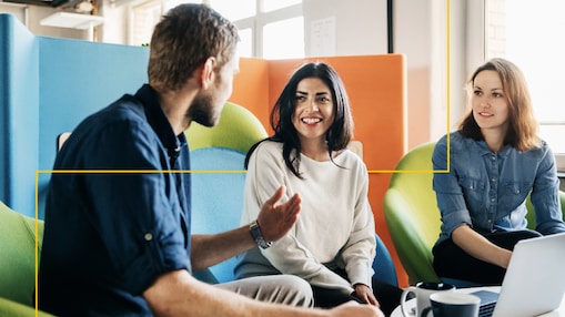 A group of three adults seated on informal office chairs discuss what is on their laptop screen in a friendly and relaxed manner