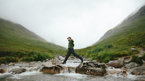 A man dressed in outdoor clothing jumps across a rocky stream, with lush grassy scottish mountains in the background and a heavily clouded sky