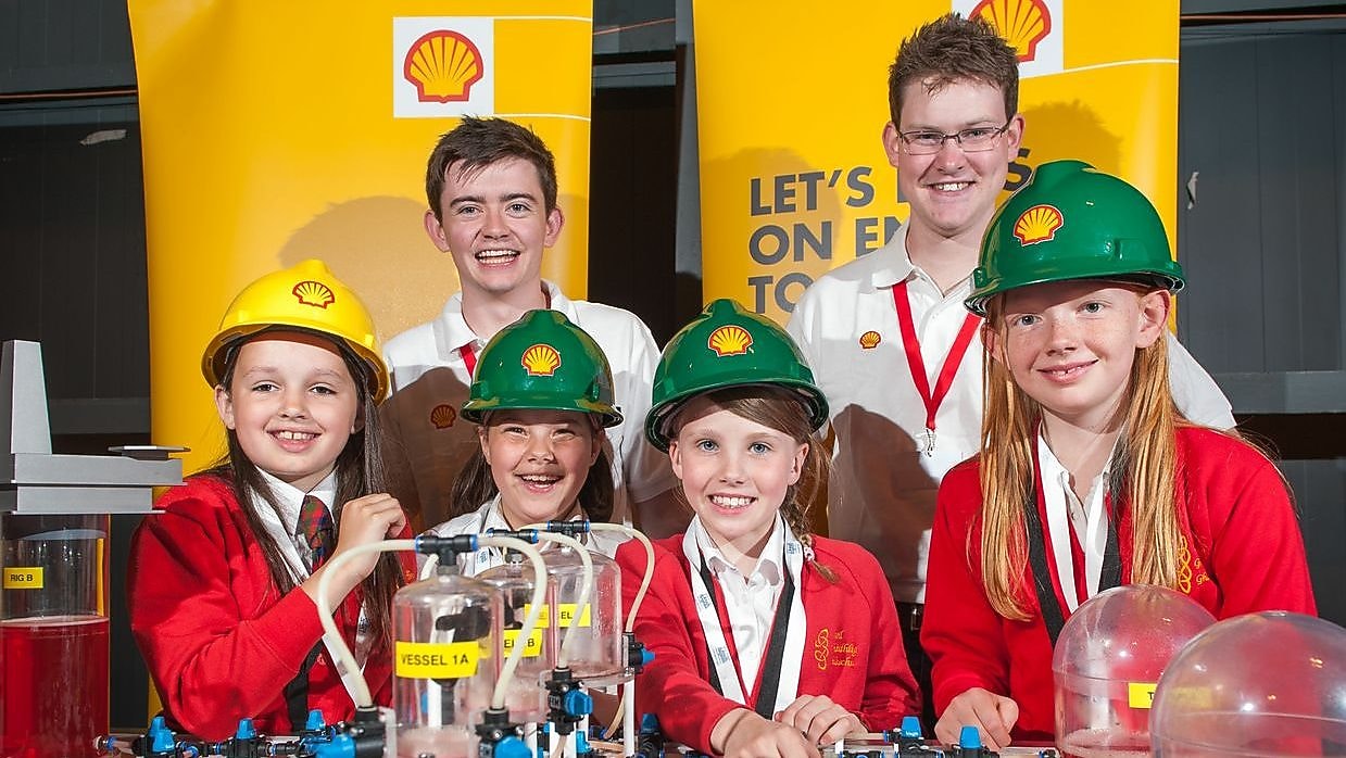 Young school children huddles smiling with Shell employees, in Shell hard-hats