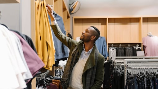 Man browses some shelves of clothes and is reaching up to inspect a yellow jumper