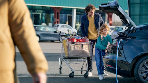 A man and his daughter putting their grocery shopping into their electric vehicle which is plugged in.