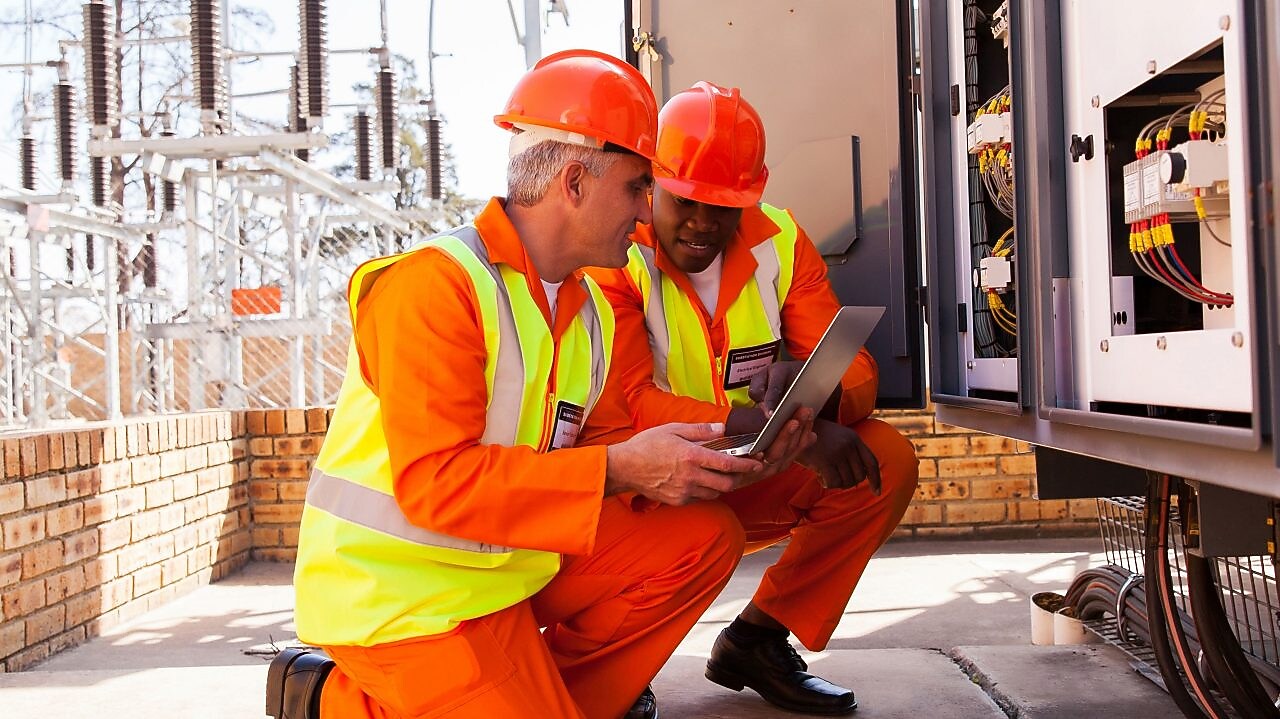 Two men wearing construction orange gaze at a laptop.