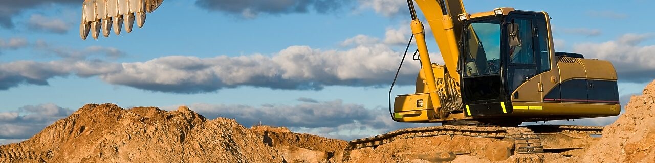 A large yellow excavator is positioned on a mound of dirt, with its arm extended and bucket raised.