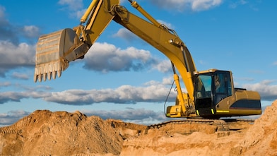 A large yellow excavator is positioned on a mound of dirt, with its arm extended and bucket raised. 