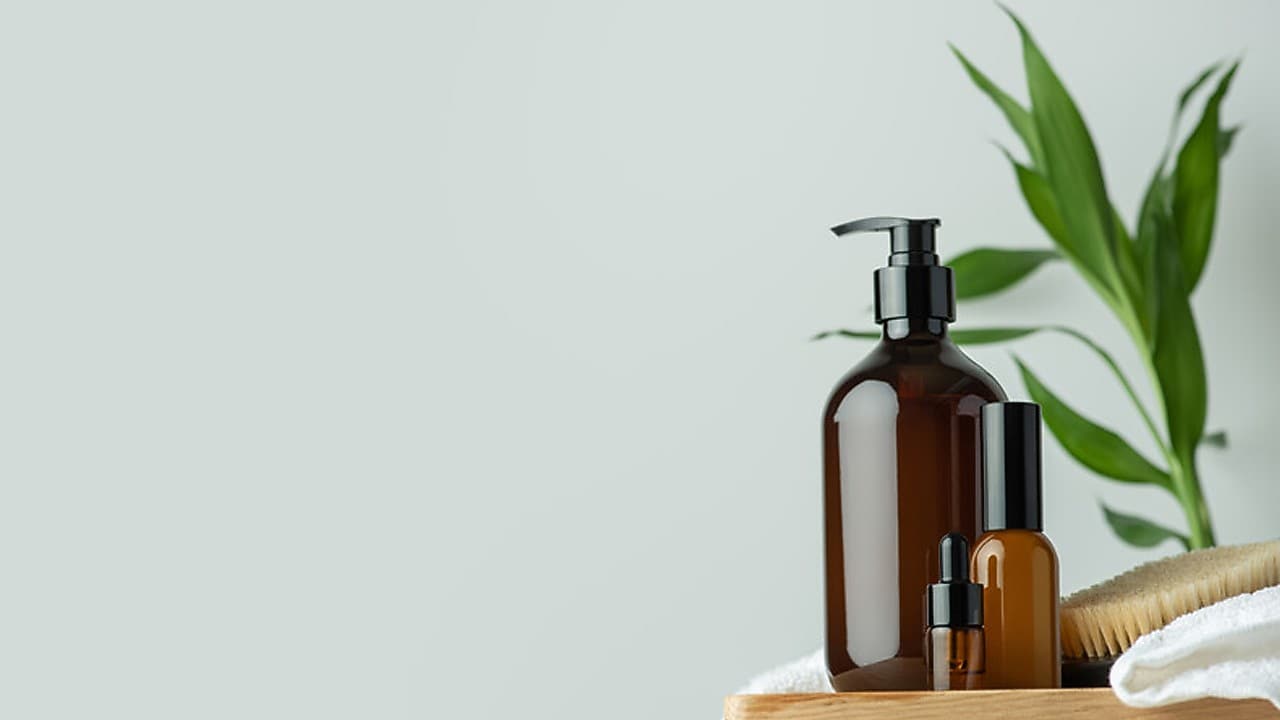 Ein Holztablett mit drei bernsteinfarbenen Flaschen in unterschiedlichen Größen, jeweils mit schwarzen Pumpaufsätzen oder Verschlüssen, daneben ein weißes Handtuch und eine Holzbürste. Im Hintergrund ist eine grüne Pflanze mit langen Blättern vor einer hellgrauen Wand zu sehen