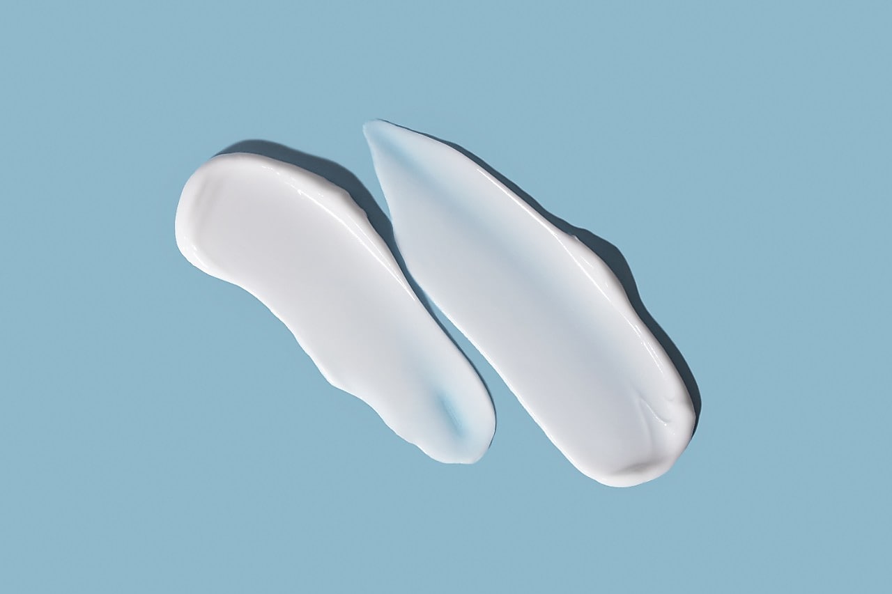 Zwei Streifen weißer Creme oder Lotion auf einem hellblauen Hintergrund. Die Streifen haben eine glatte, glänzende Textur; der linke ist kleiner und gebogen, der rechte größer, länglicher und zeigt am oberen Rand einen leicht bläulichen Farbton.