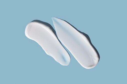 Zwei Streifen weißer Creme oder Lotion auf einem hellblauen Hintergrund. Die Streifen haben eine glatte, glänzende Textur; der linke ist kleiner und gebogen, der rechte größer, länglicher und zeigt am oberen Rand einen leicht bläulichen Farbton. 